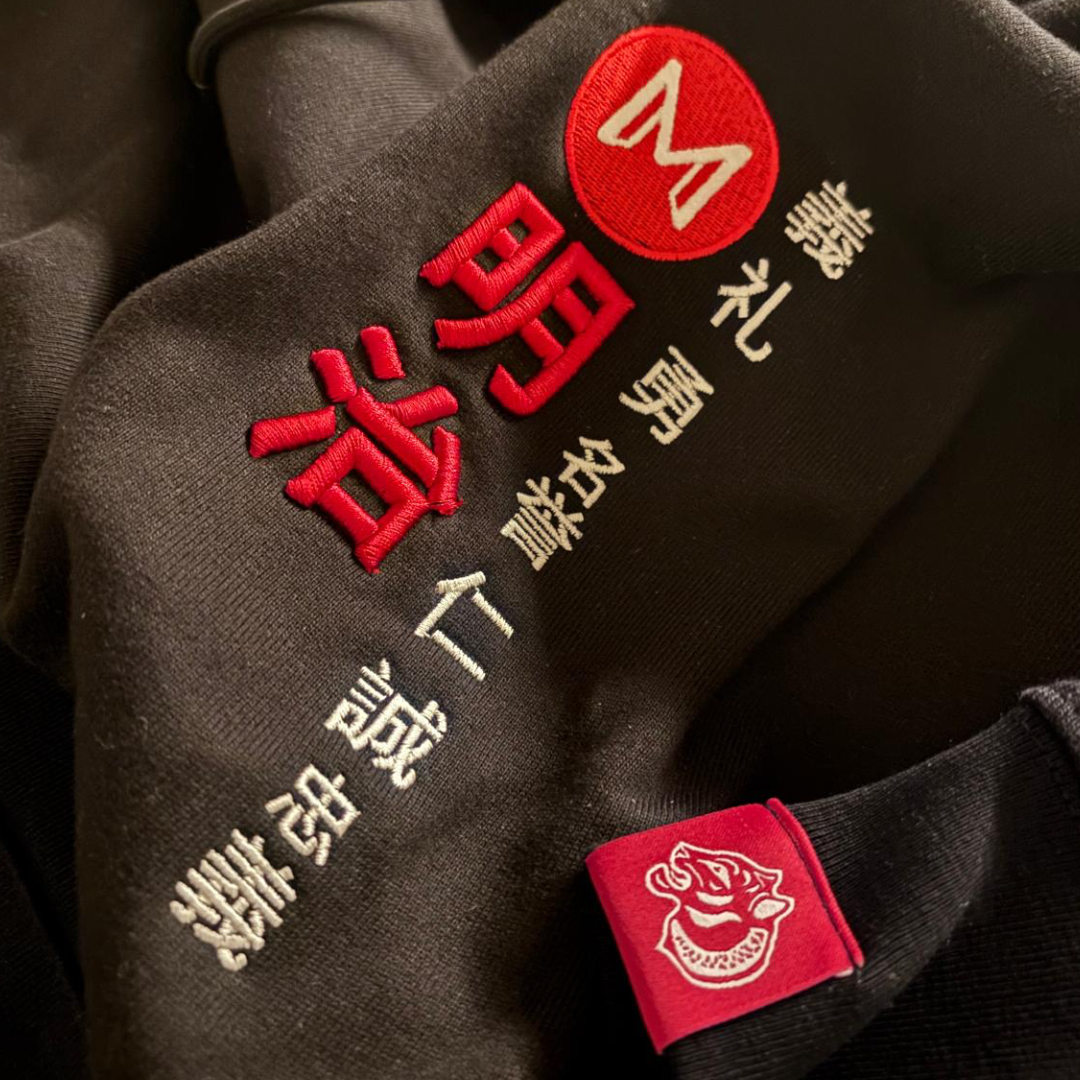 Bushido hoodie detailing.Sleeve close-up: Embroidered tiger tab on left sleeve.
Fabric texture: Close-up of heavyweight 350GSM organic fleece cotton in matte black and embroidery detailing 
