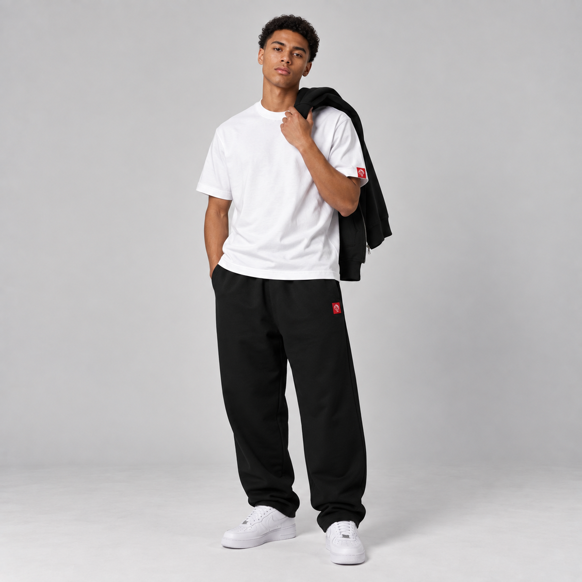 Model wearing the FormaFinalis FF Capsule white heavyweight organic cotton T-shirt and black relaxed cuffless fleece joggers, carrying a black heavyweight zip hoodie with a red embroidered FormaFinalis logo over one shoulde